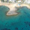 Veduta aerea di una costa rocciosa con acque blu e alcuni edifici sullo sfondo.