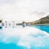 Una piscina a sfioro riflette il cielo, circondata da colline con edifici bianchi.