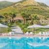 Piscina con lettini e ombrelloni bianchi, circondata da palme e colline verdi sullo sfondo.