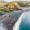 Spiaggia di ciottoli con file di ombrelloni, mare azzurro e colline verdi sullo sfondo.