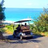 Persone in golf cart su una strada con vegetazione che conduce a una spiaggia e mare blu.