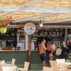 Un bar all'aperto con tetto di canne, persone al bancone e fiori appesi. Sedie di plastica beige.