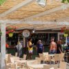 Bar all'aperto con tetto di canne, persone che socializzano, sedie beige e fiori appesi.