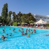 Persone in una grande piscina circondata da ombrelloni e alberi sotto un cielo sereno.