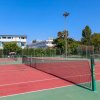 Un singolo campo da tennis con rete, circondato da alberi e edifici. Il cielo è sereno.