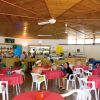 Interno di un bar con tavoli rotondi e sedie in plastica bianche, persone sedute che chiacchierano.