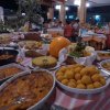 Tavolo buffet con vari piatti, tra cui arancini e verdure, persone sullo sfondo.