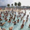 Persone in piscina affollata con palme, sotto un cielo nuvoloso.