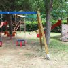 Parco giochi con altalene, scivolo e parete da arrampicata tra alberi.