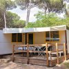 Casa mobile con terrazza in legno, tavolo, sedie, tende. Circondata da alberi alti e vegetazione.