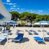 Lettini blu e ombrelloni bianchi su una spiaggia sabbiosa, alberi sullo sfondo.