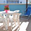 Tavolo bianco con sedie e pianta rossa su terrazza, sedie a sdraio blu sullo sfondo.
