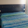 Letto matrimoniale con copriletto blu, testiera in legno, tenda marrone e piccola mensola.