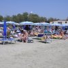 Spiaggia affollata con lettini blu e ombrelloni bianchi, persone che prendono il sole.