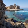 Vista di un antico castello sul mare con acqua cristallina e rocce in primo piano.