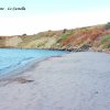 Spiaggia sabbiosa con rocce, vegetazione e colline in lontananza.