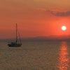 Barca a vela su mare calmo al tramonto, con sole arancione e montagne sullo sfondo.