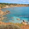 Baia rocciosa con mare turchese, vegetazione secca e un piccolo sentiero.