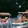 Due persone prendono il sole su lettini vicino a una piscina, con piante sullo sfondo.