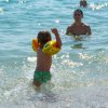 Bambino con braccioli arancioni entra nell'acqua, mentre un adulto lo osserva sorridendo.