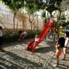 Bambini giocano al parco su uno scivolo e con una palla, circondati da alberi.