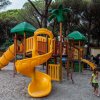 Parco giochi con scivoli gialli e verdi. Bambini che giocano nella sabbia, alberi intorno.