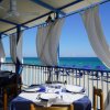 Tavoli apparecchiati con vista sul mare, tende bianche sventolano sotto una tettoia azzurra.