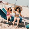 Due bambine in costume da bagno salutano sulla spiaggia, sedute su lettini con cappelli estivi.