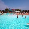 Piscina affollata con persone che nuotano e si rilassano, ombrelloni bianchi sullo sfondo.